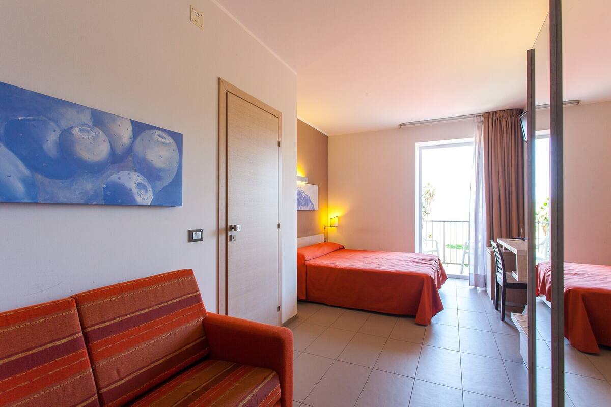 A hotel room with a sofa, two single beds, a desk, and a painting on the wall.