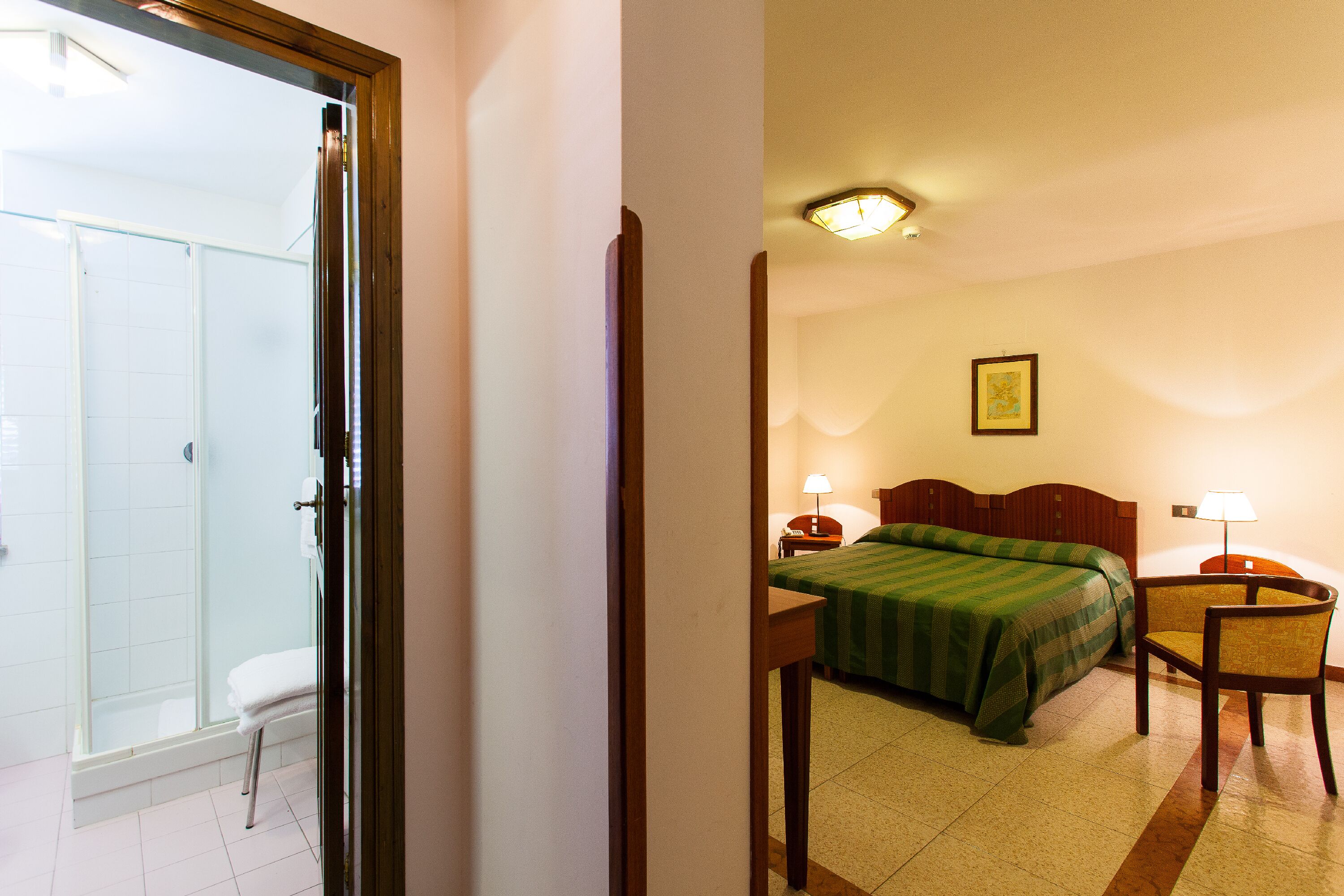 Habitación de hotel con cama, una silla, mesita de noche, lámpara y zona de ducha.