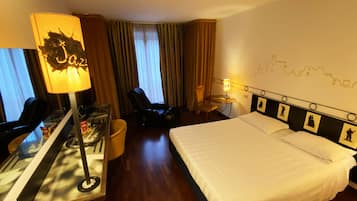 A hotel room with a bed, a desk with a lamp, a chair, a small table, and a wall with a cityscape mural.