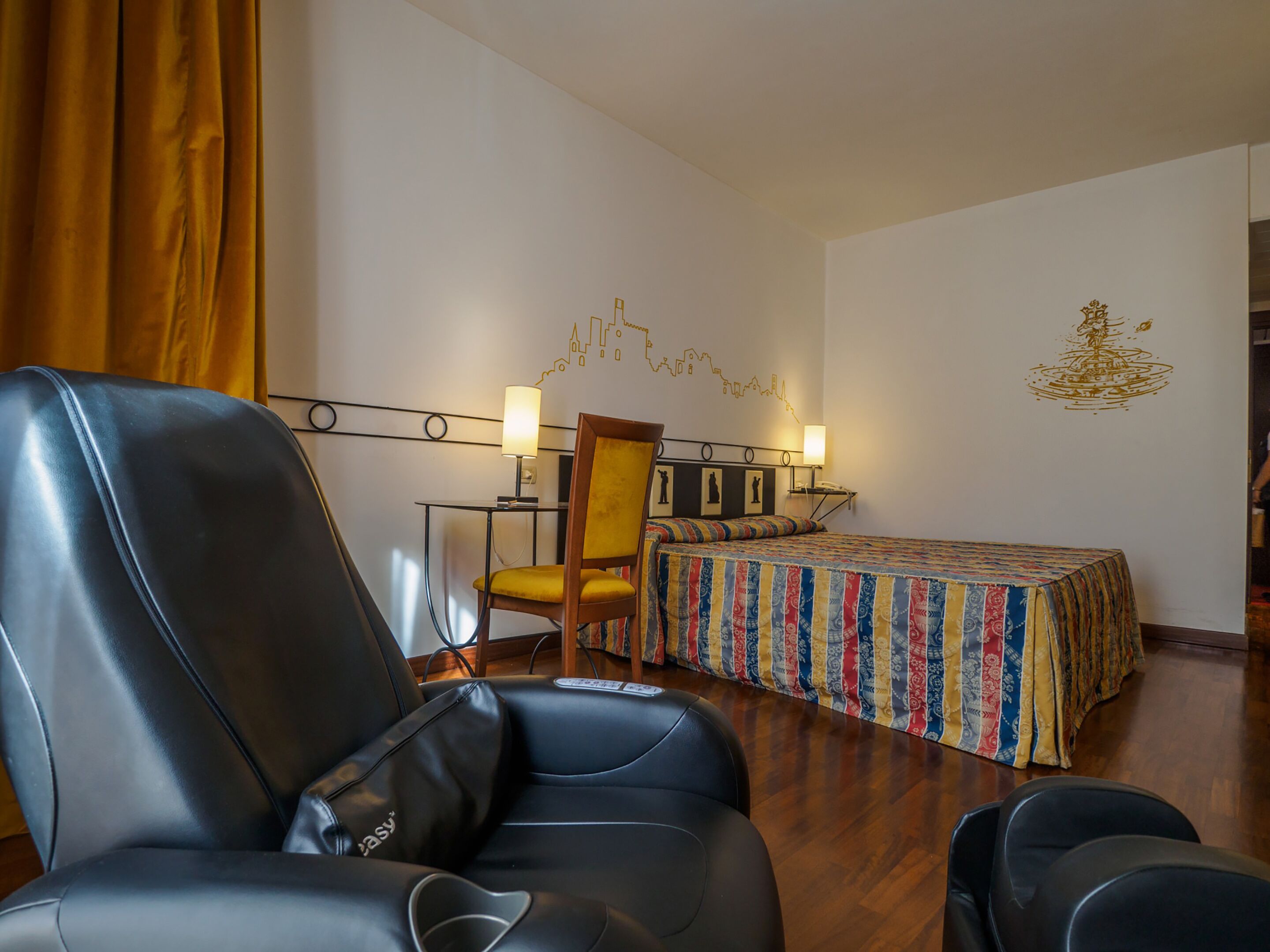 Una habitación de hotel con una butaca de cuero negro, una cama con colcha de colores, un escritorio con dos lámparas y una obra de arte en la pared.