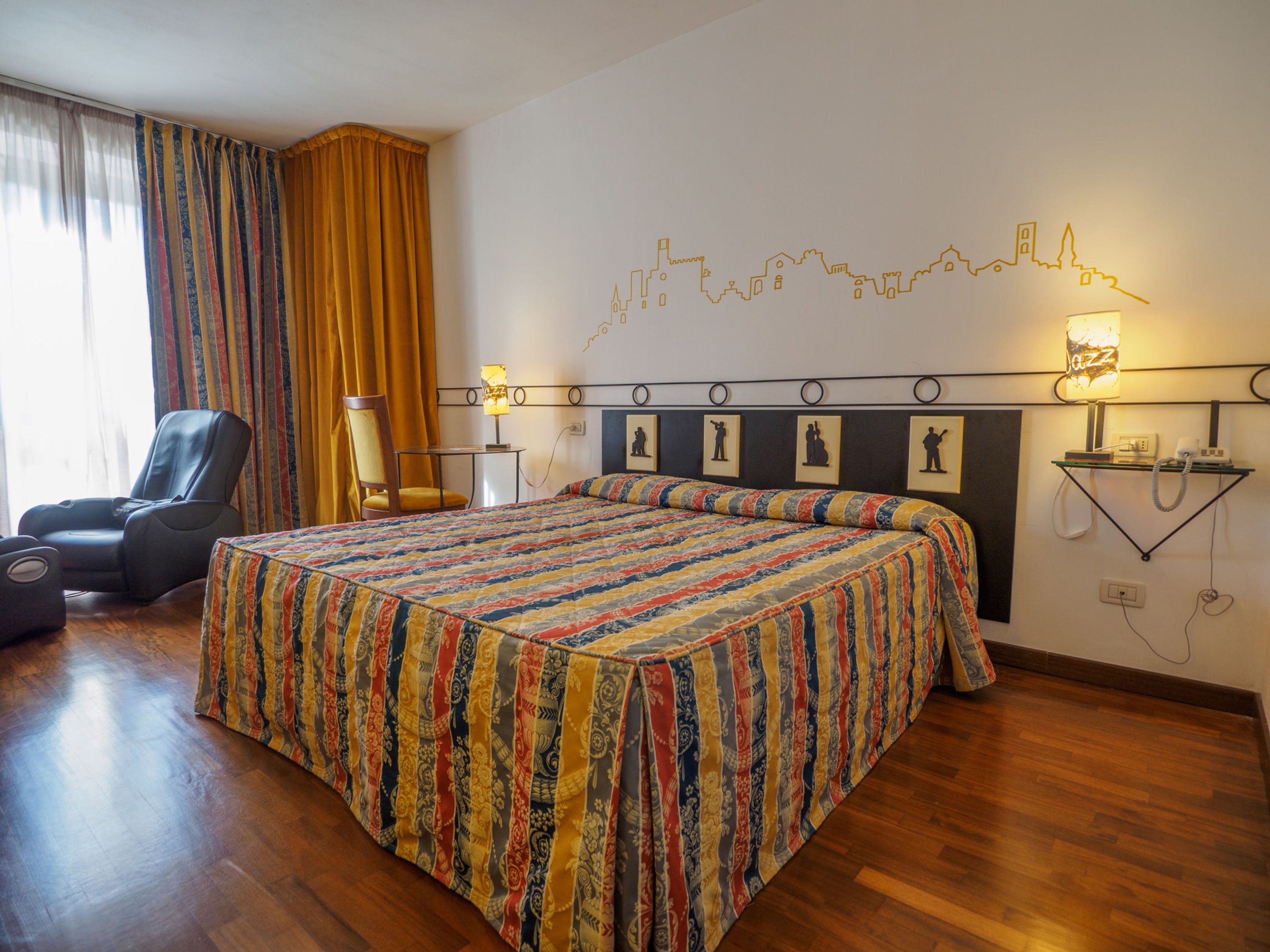 Een hotelkamer met een groot bed, een stoel, een bureau en een muur met een stadsgezicht kunstwerk.