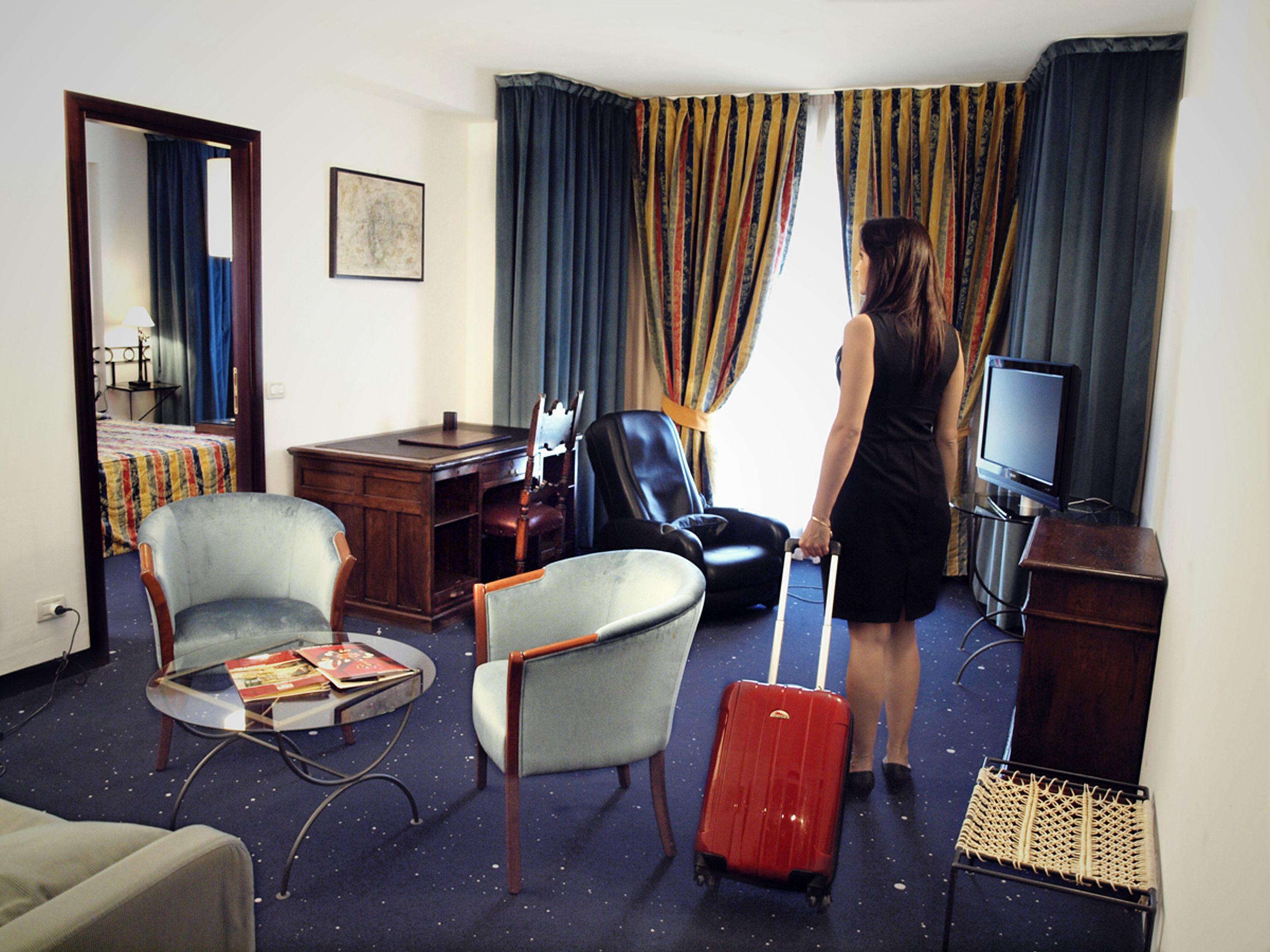 A hotel room with a bed, a desk, a television, a chair, a suitcase, and a window with curtains.