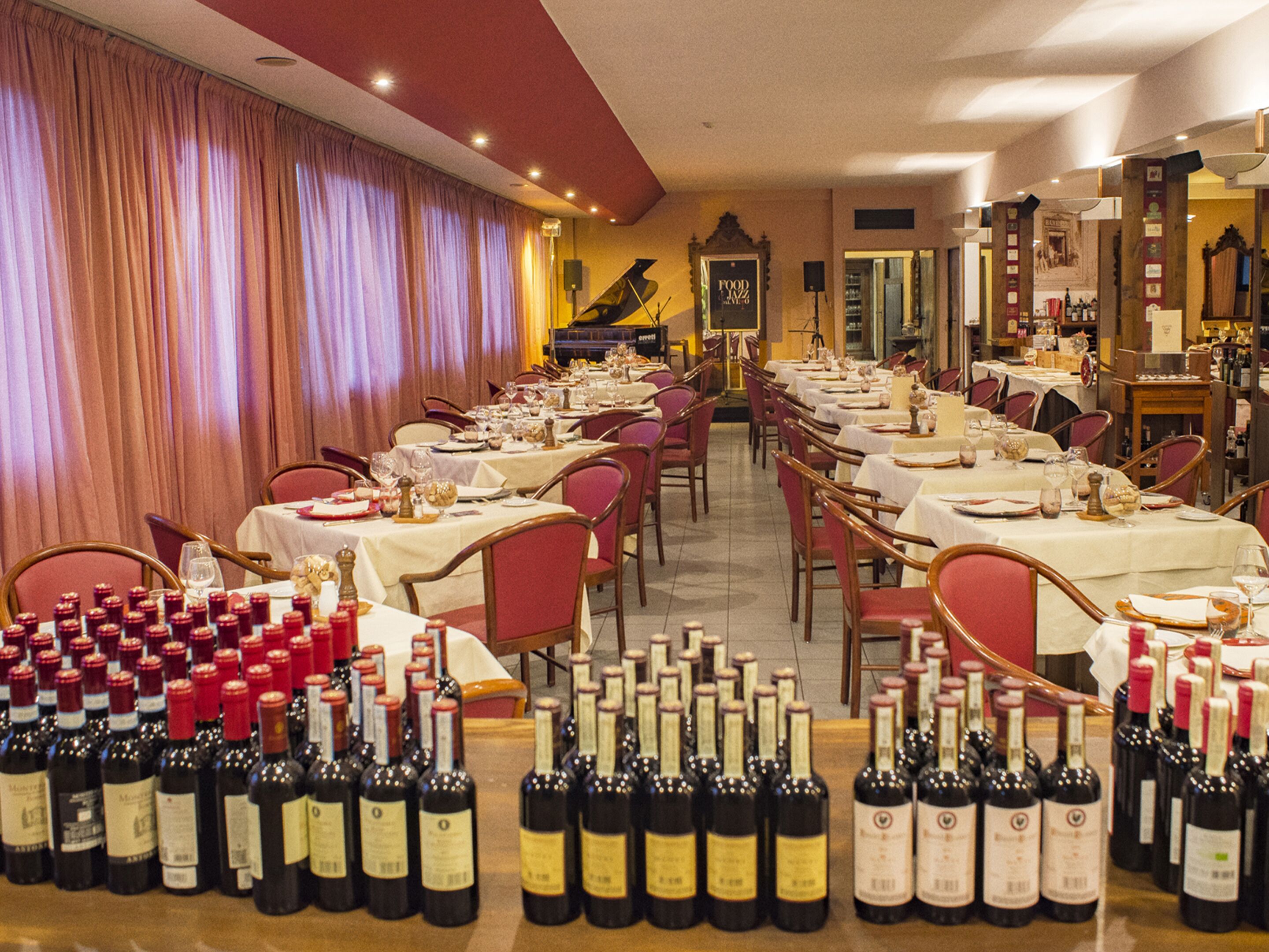 A restaurant with multiple tables set for dining, a piano in the background, and numerous wine bottles displayed in the foreground.