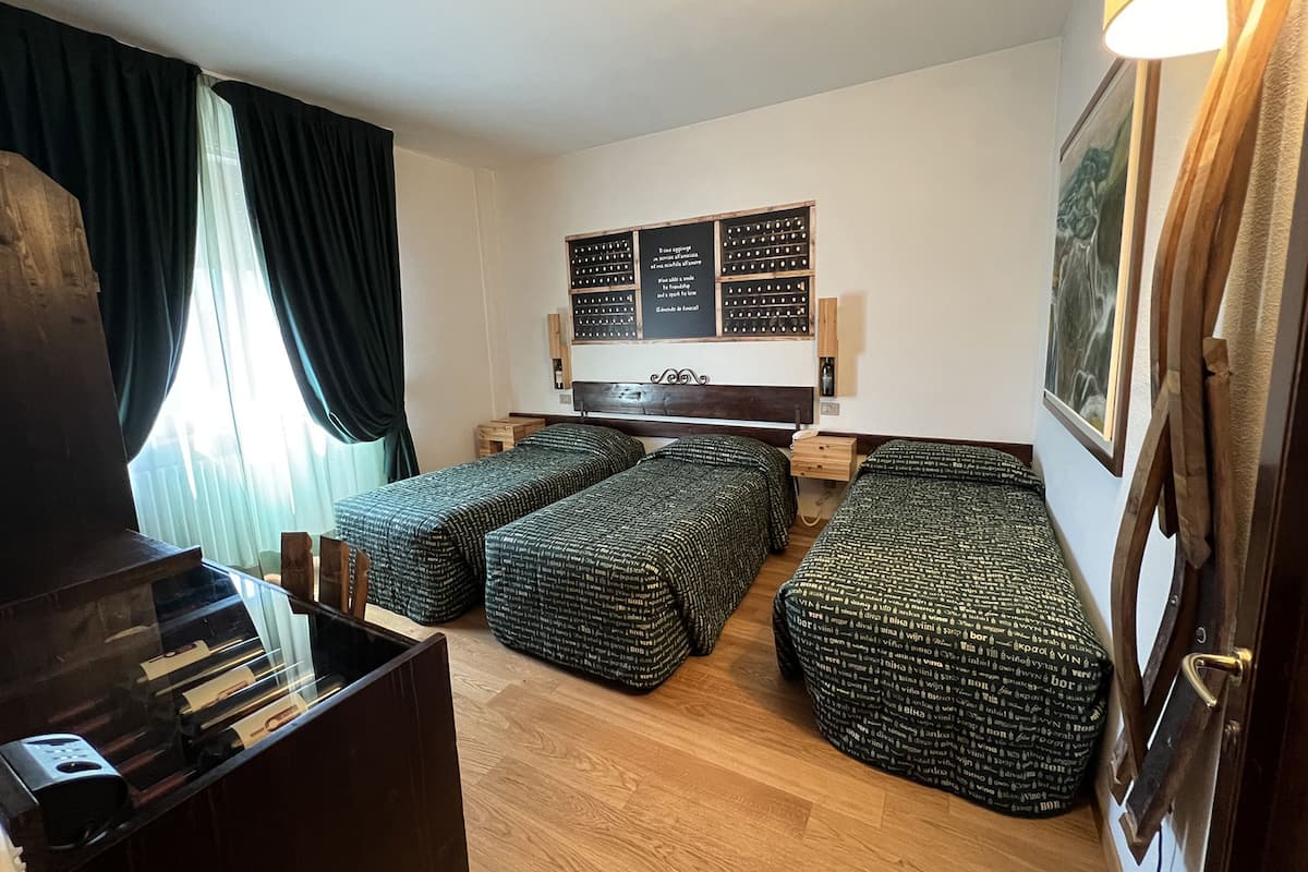 A small, cozy room with two single beds, a wooden desk, and a small table. There is a window with curtains, a wall-mounted TV, and a framed picture.