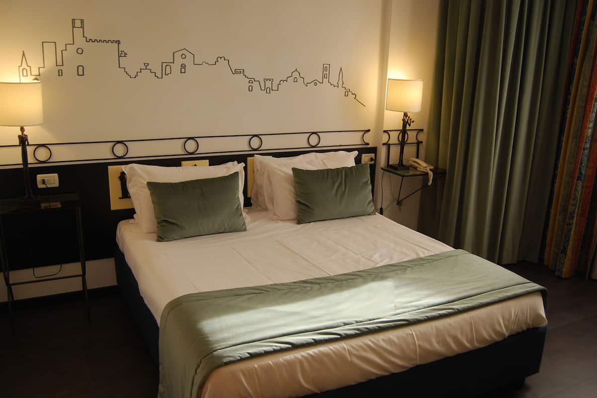 A hotel room with a bed, two bedside tables, a wall art of a city skyline, and a curtain.