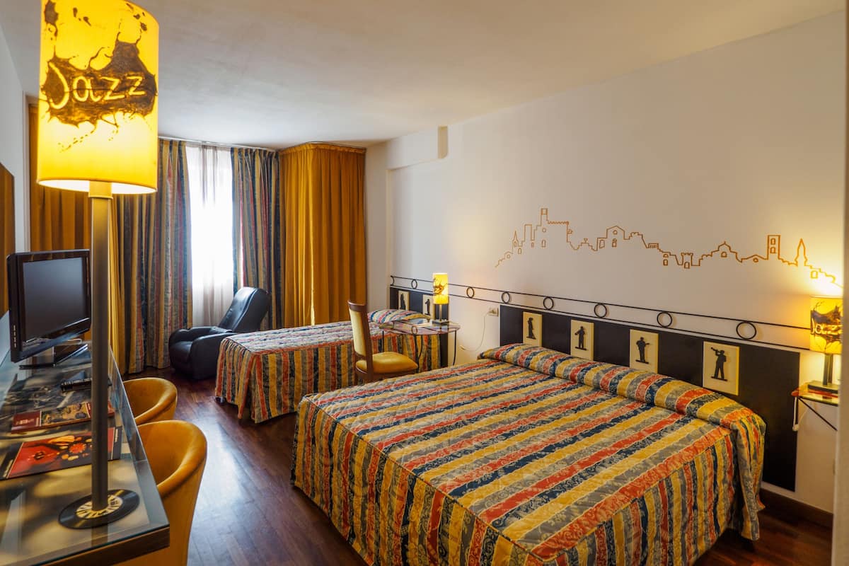 A hotel room with two beds, a desk with a computer, a chair, and a lamp labeled "Jazz".