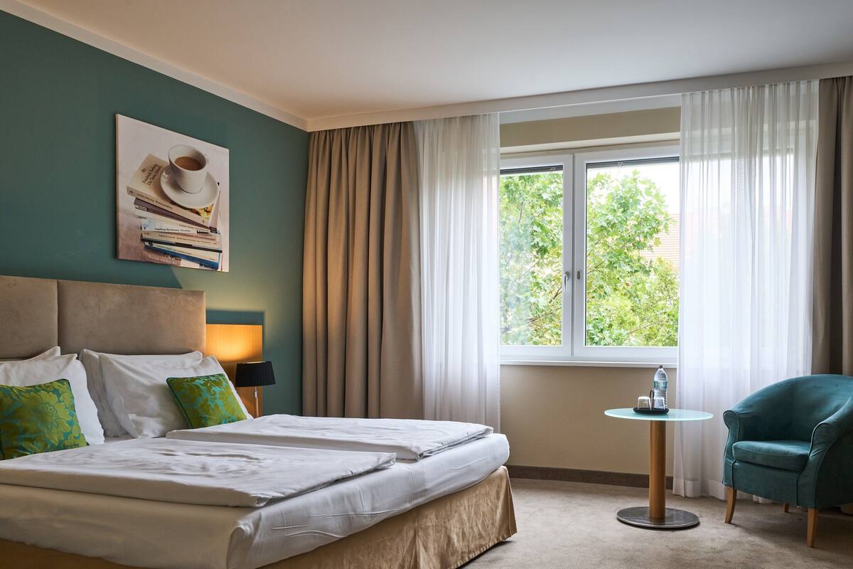 A hotel room with a bed, a small table, a chair, a lamp, and a window with curtains.