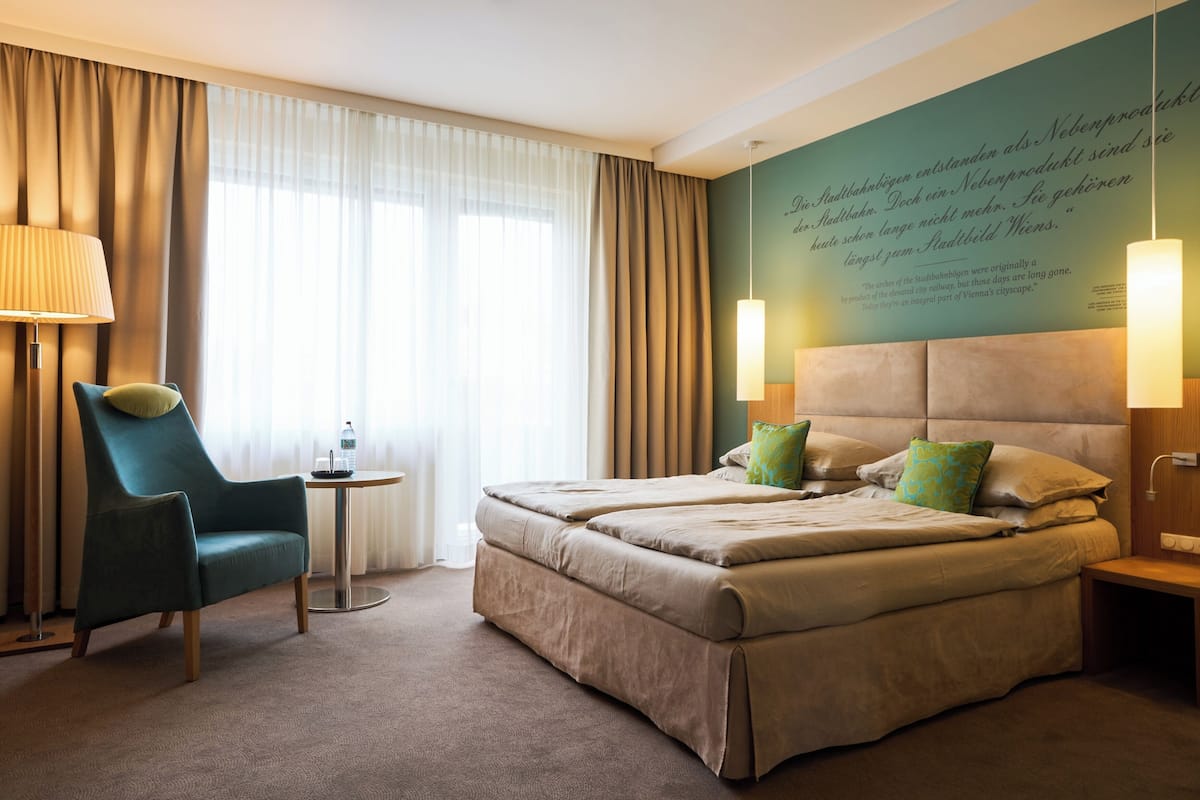 A hotel room with a large bed, a chair, a small table, and a wall with a quote.