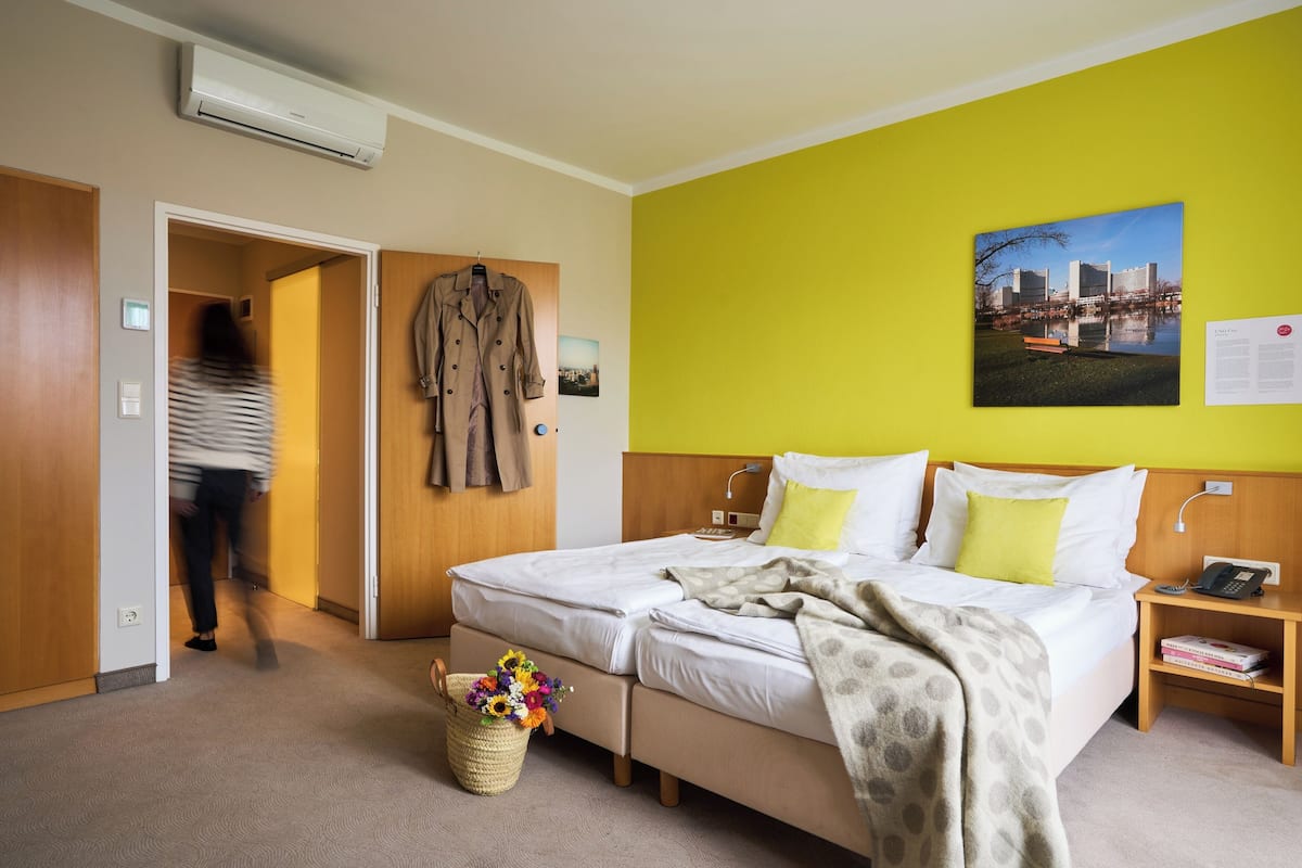 A hotel room with a bed, a nightstand, a lamp, a phone, a picture on the wall, and a window with a view of buildings.