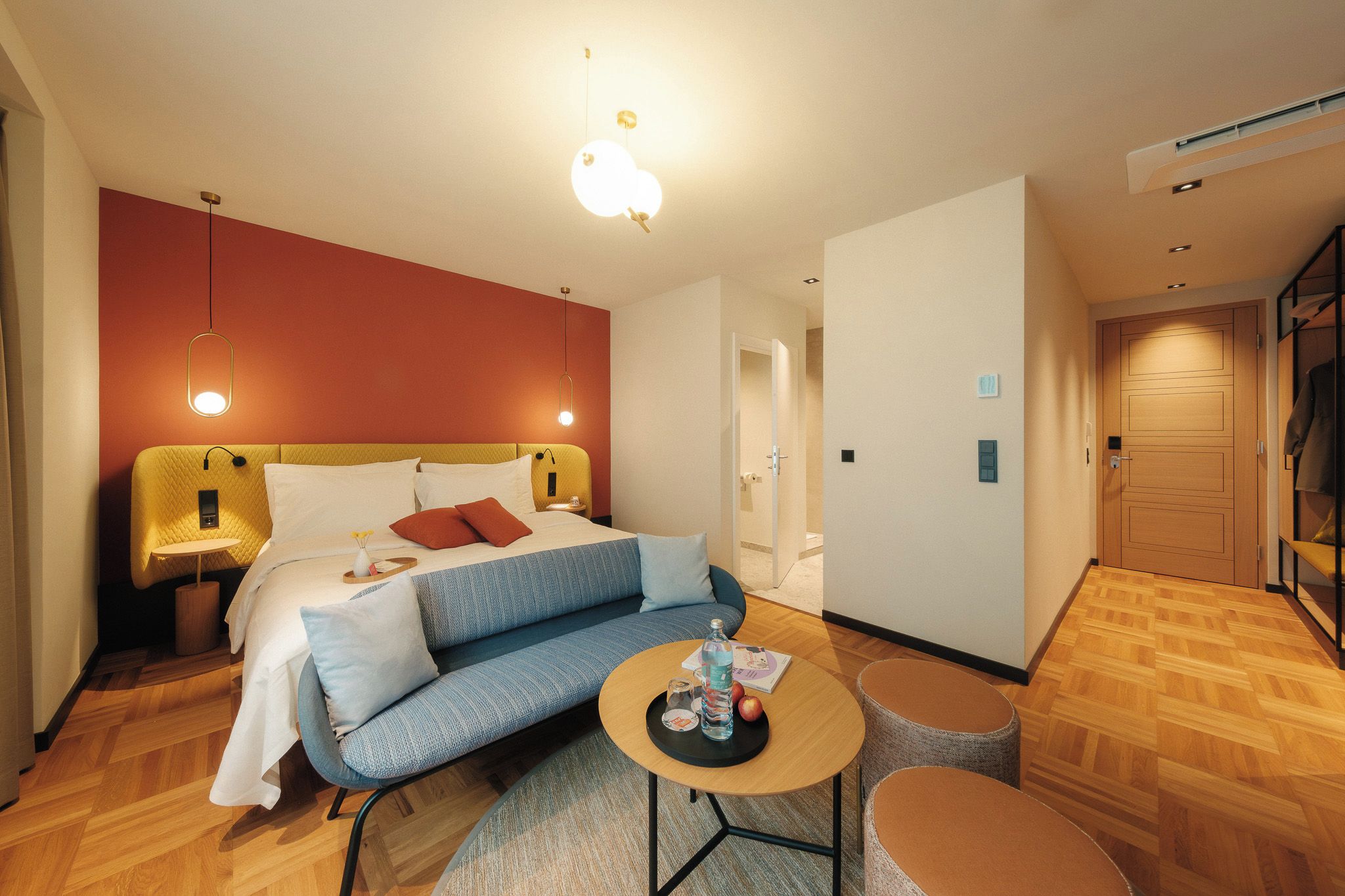 Quarto de hotel com cama, sofá, uma mesa pequena e porta que dá para outro cômodo.