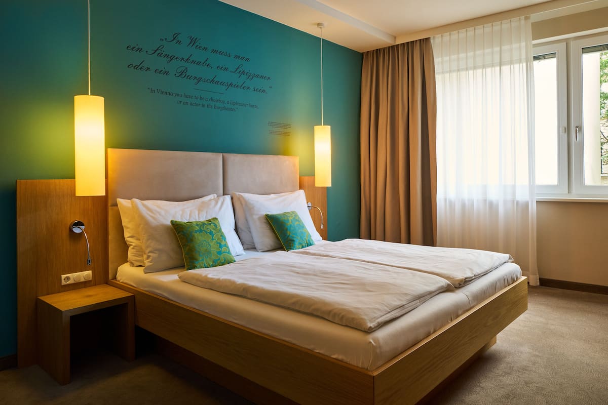 A neatly made bed with white linens and two pillows, a bedside table with a lamp, and a wall with a quote in a foreign language.