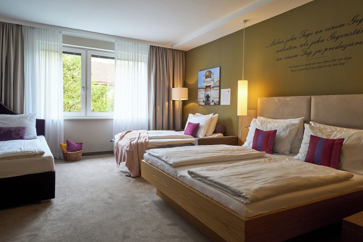 A hotel room with two beds, a large window with curtains, a wall with a quote, and a lamp.