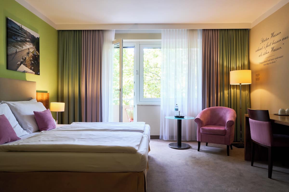 A hotel room with two beds, a desk, a chair, a lamp, and a view of the outdoors.