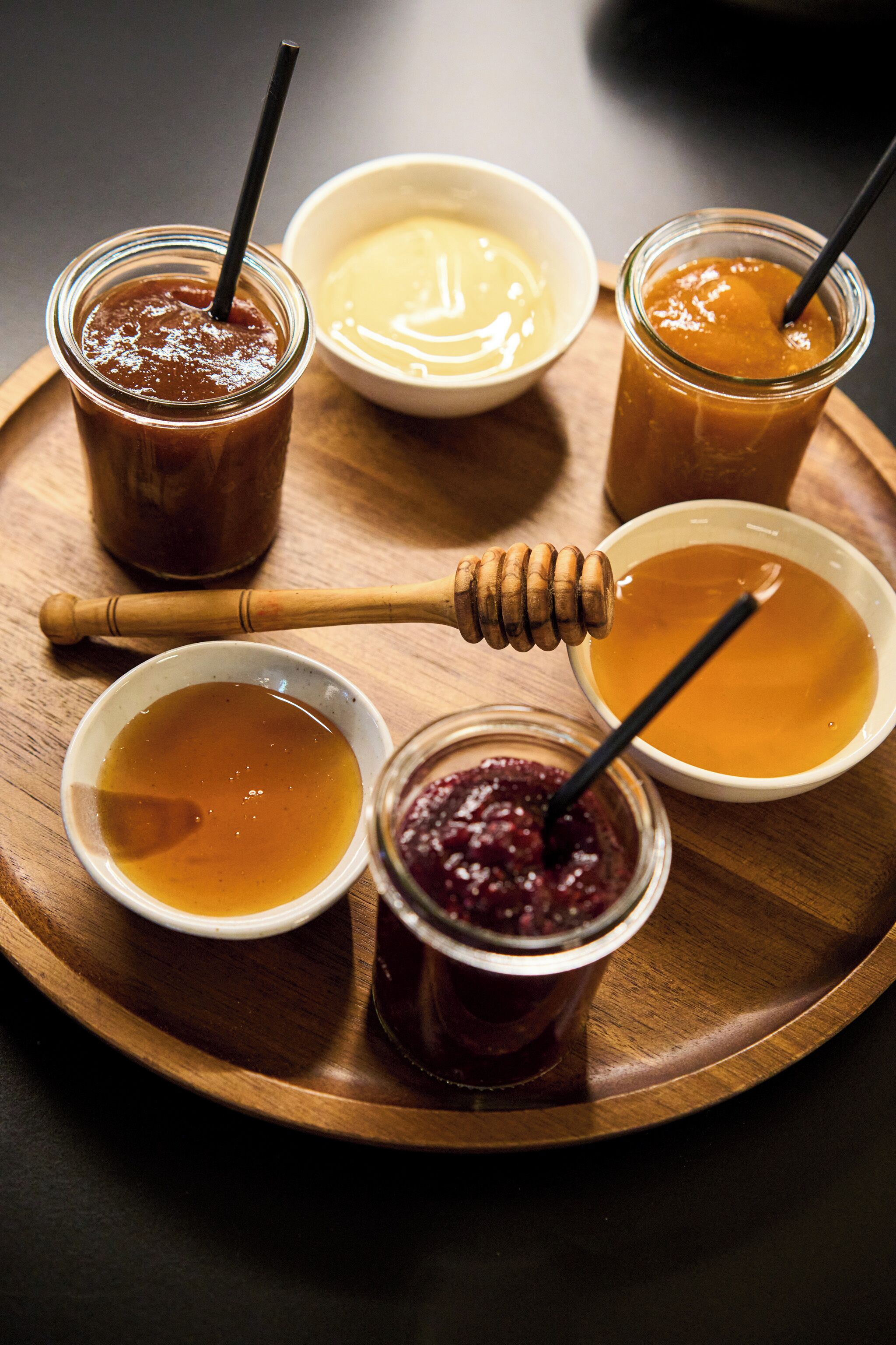 Un plateau en bois contenant divers pots de miel et de sirop, dont certains sont accompagnés d’ustensiles pour prélever le miel.
