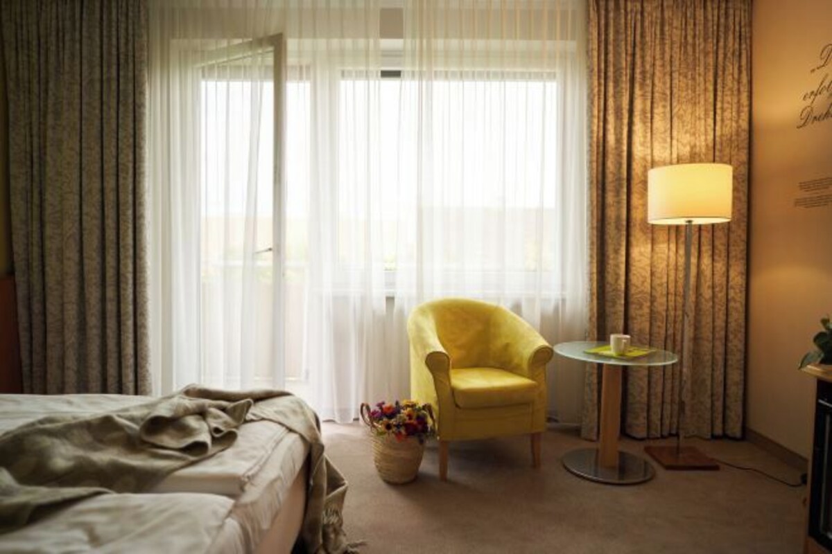 A hotel room with a bed, a yellow armchair, a small round table with a lamp, and a window with sheer curtains.