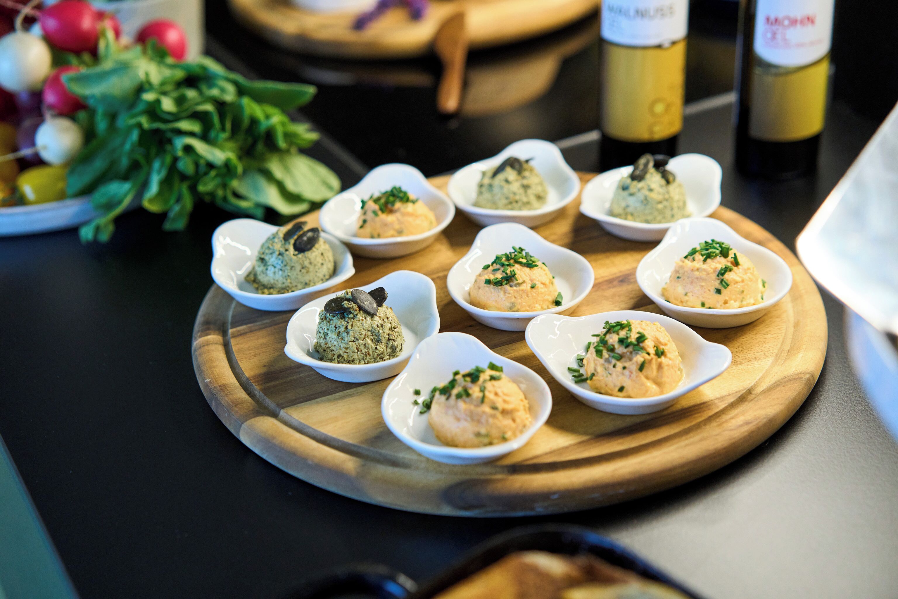 Ein Holzboden mit kleinen weißen Schalen, die Lebensmittel enthalten, ein Schneidebrett und im Hintergrund Weinflaschen.