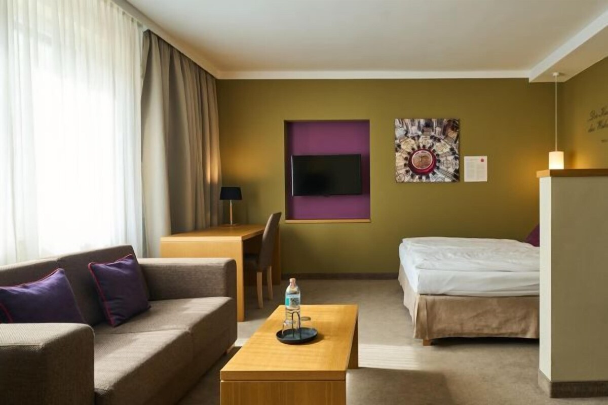 Una habitación de hotel con sofá, cama, mesa de centro, televisor y una obra de arte en la pared.