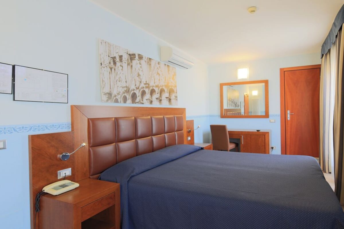 Ein Hotelzimmer mit einem großen Bett, einem Nachttisch aus Holz mit einem Telefon, einem Schreibtisch mit Stuhl, einem Spiegel und einer Tür mit gestreiften Vorhängen.