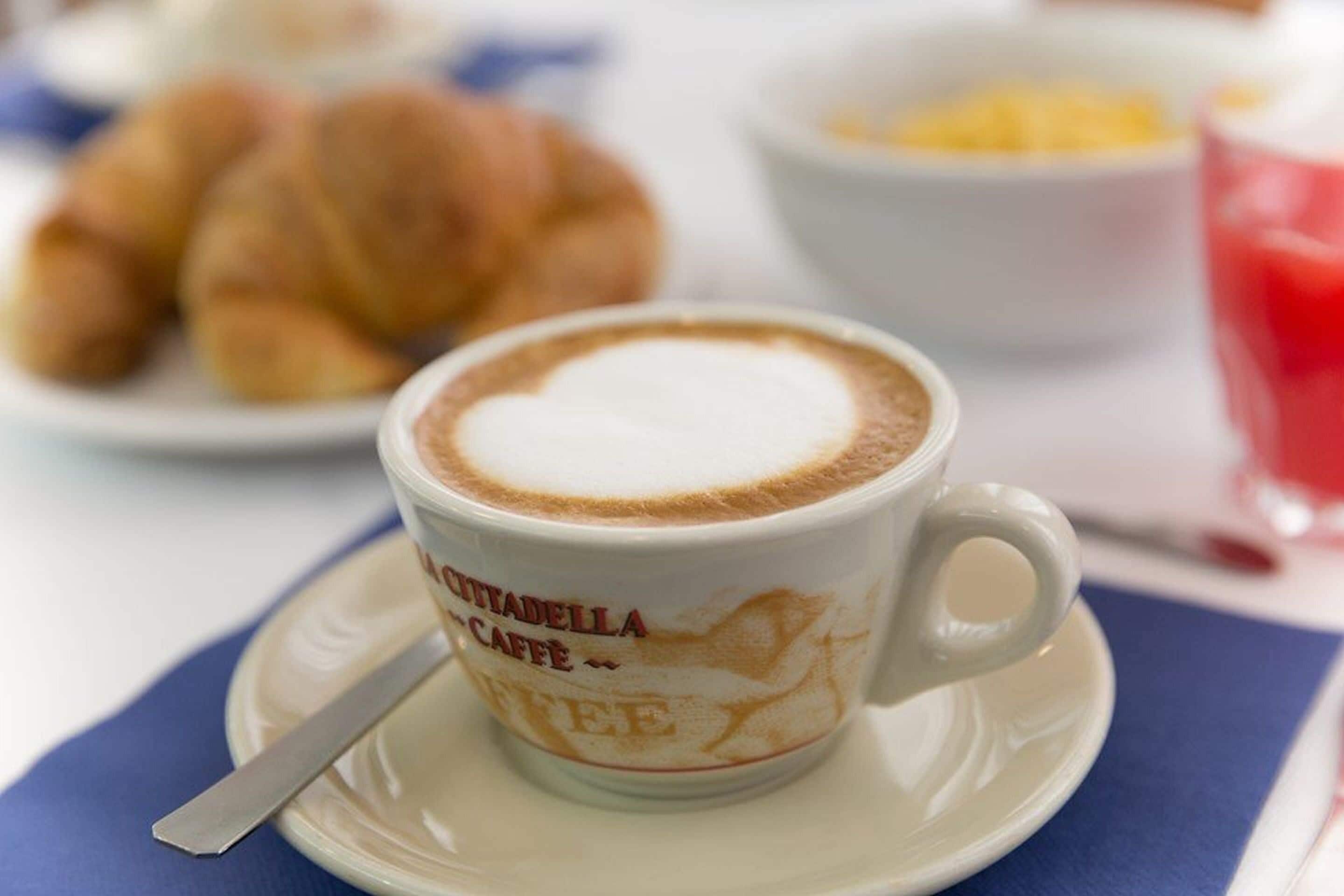 A cup of cappuccino with latte art on a saucer, a spoon, and a plate with croissants in the background.