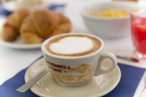 A cup of cappuccino with latte art on a saucer, a spoon, and a plate with croissants in the background. - Hotel Terminus & Plaza (Pisa)