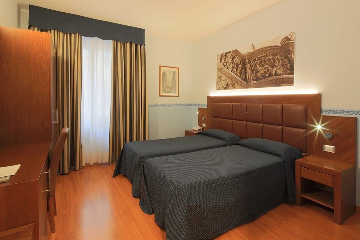 Ein Hotelzimmer mit zwei Betten, einem Schreibtisch, einem Stuhl, einem Nachttisch und einer Wandleuchte.