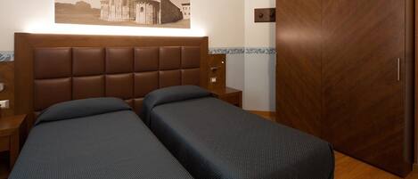 Habitación de hotel con dos camas individuales, un armario de madera, una mesita de noche y una lámpara fijada a la pared.