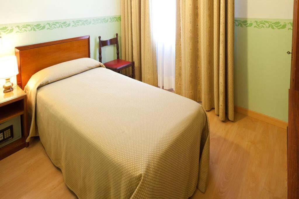 Una cama bien hecha con cabecera de madera, una silla, una mesita de noche con lámpara y una puerta con cortina.