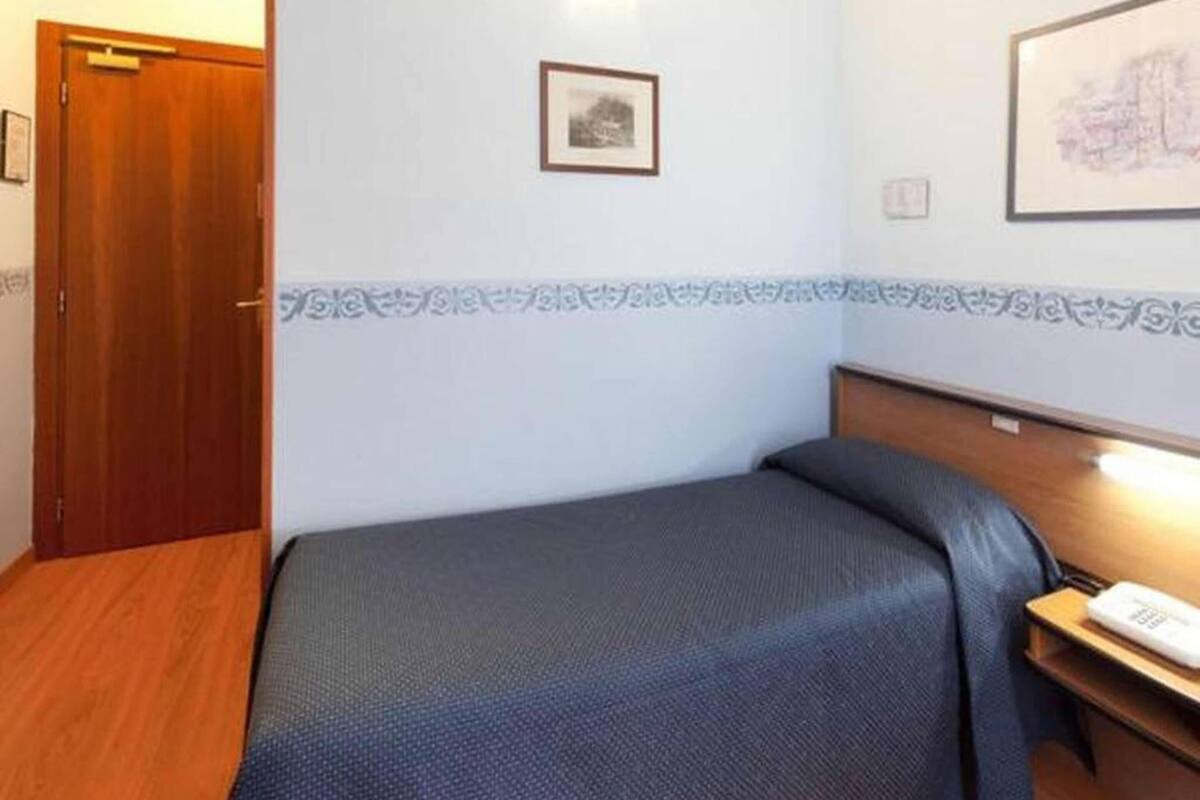 Ein kleines, ordentliches Hotelzimmer mit einem Einzelbett, einem Nachttisch und einem gerahmten Bild an der Wand.