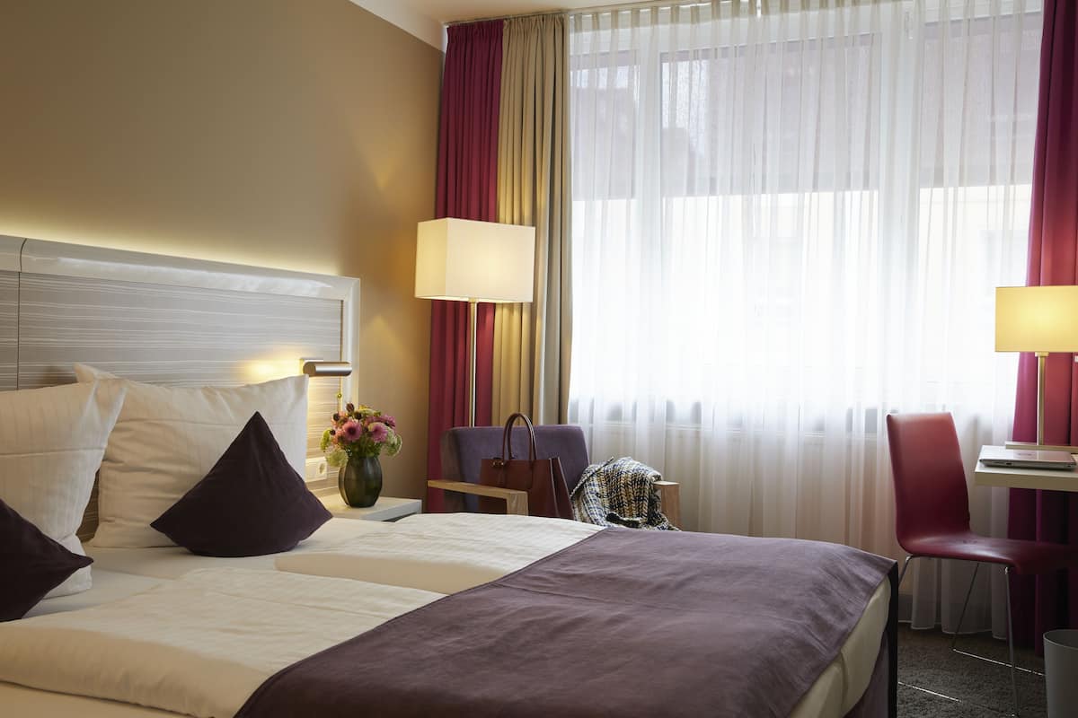 Una camera d'albergo con un letto, due comodini, una scrivania, una sedia e una finestra con tende.