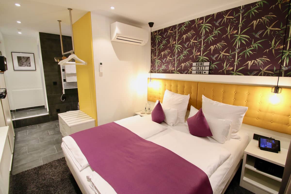 Una camera d'albergo moderna con un letto grande, un comodino con sveglia digitale, un condizionatore d'aria a parete e un portasciugamani.