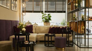 Un salon moderne avec un canapé beige, des coussins violets et des tables d’appoint noires. On y trouve des plantes en pot et un vase rempli de fleurs posé sur le canapé.