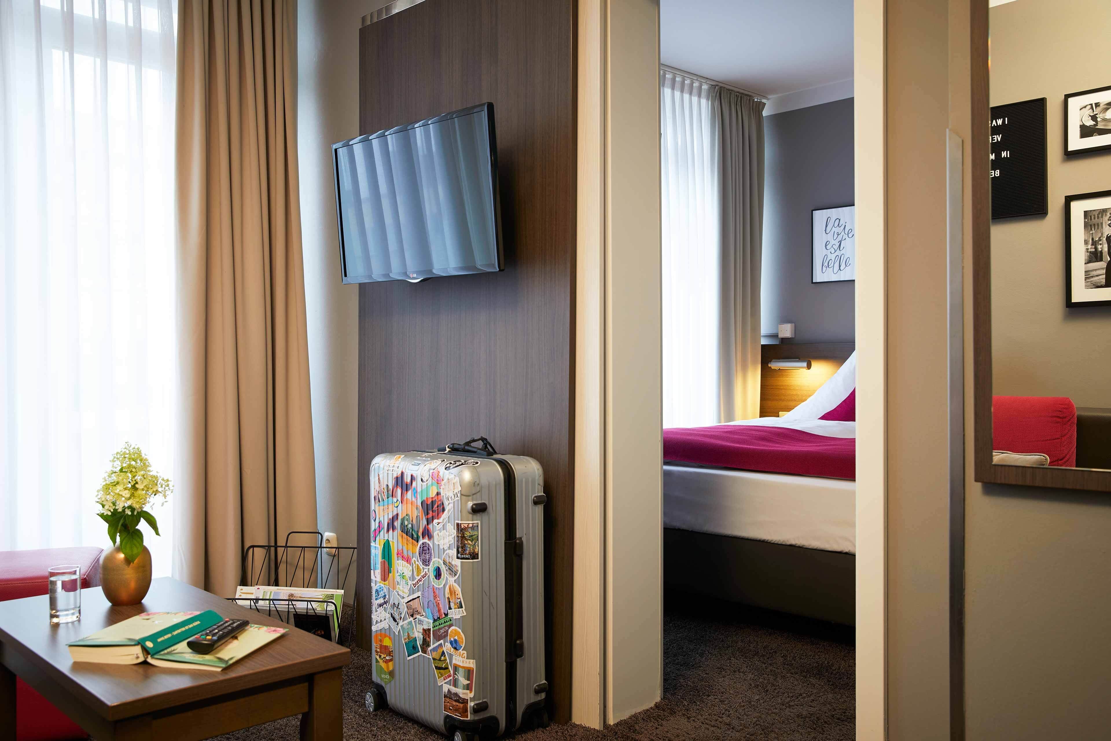 A hotel room with a bed, a desk with a book and a vase of flowers, a suitcase, and a mounted TV.