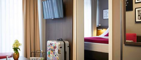 A hotel room with a bed, a desk with a book and a vase of flowers, a suitcase, and a mounted TV.