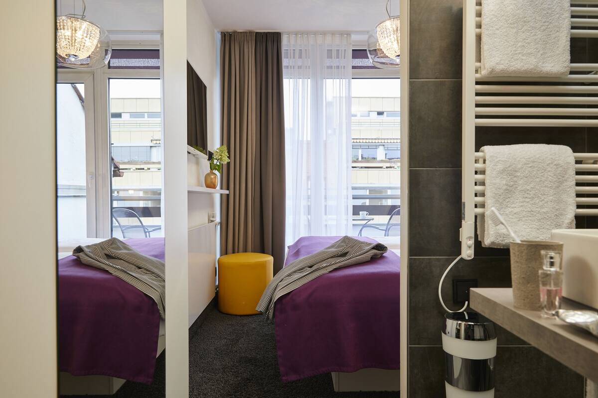 Camera d'albergo con due letti, un poggiapiedi giallo, un bagno con porta asciugamani e vista sugli edifici.