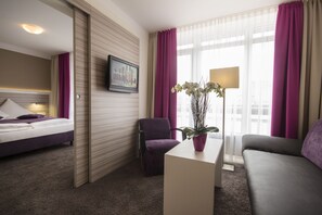 Una habitación de hotel con cama, sillón, sofá, escritorio, lámpara y ventana con cortinas.