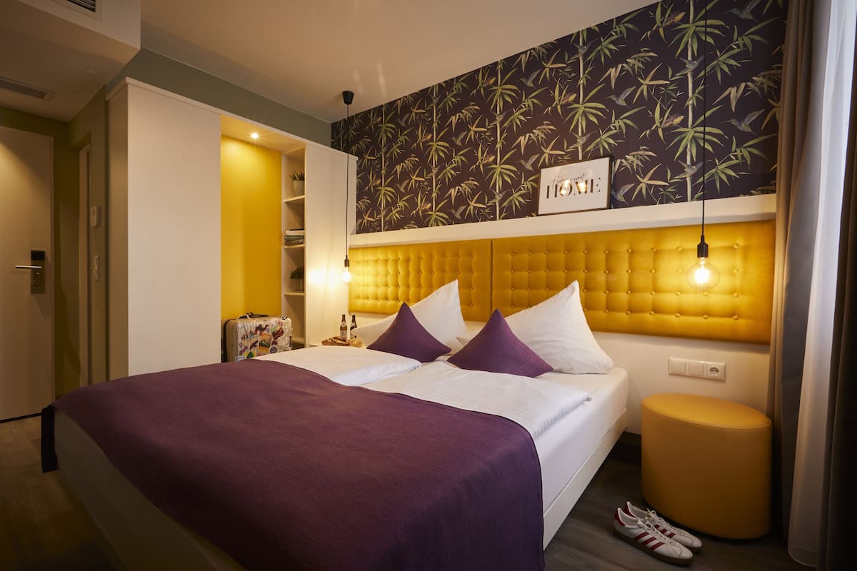 Una camera d'albergo con un letto, una testiera gialla, una parete decorata con un motivo a foglia di bambù e un piccolo sgabello.
