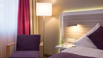 Ein Hotelzimmer mit Bett, Sessel, Nachttisch mit Lampe und Fenster mit Vorhängen.