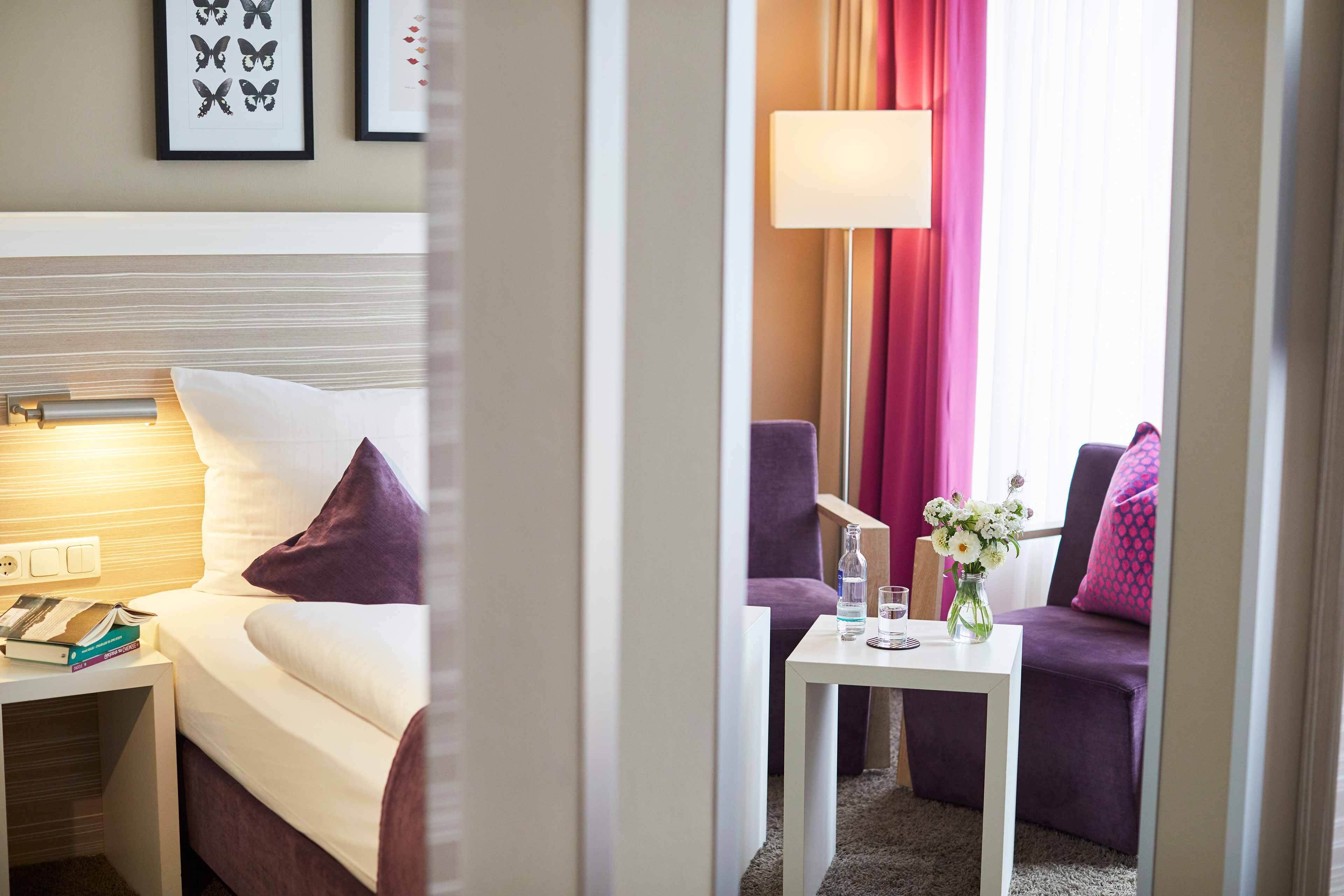 A hotel room with a bed, bedside table, a small table with a vase of flowers, and a sofa.