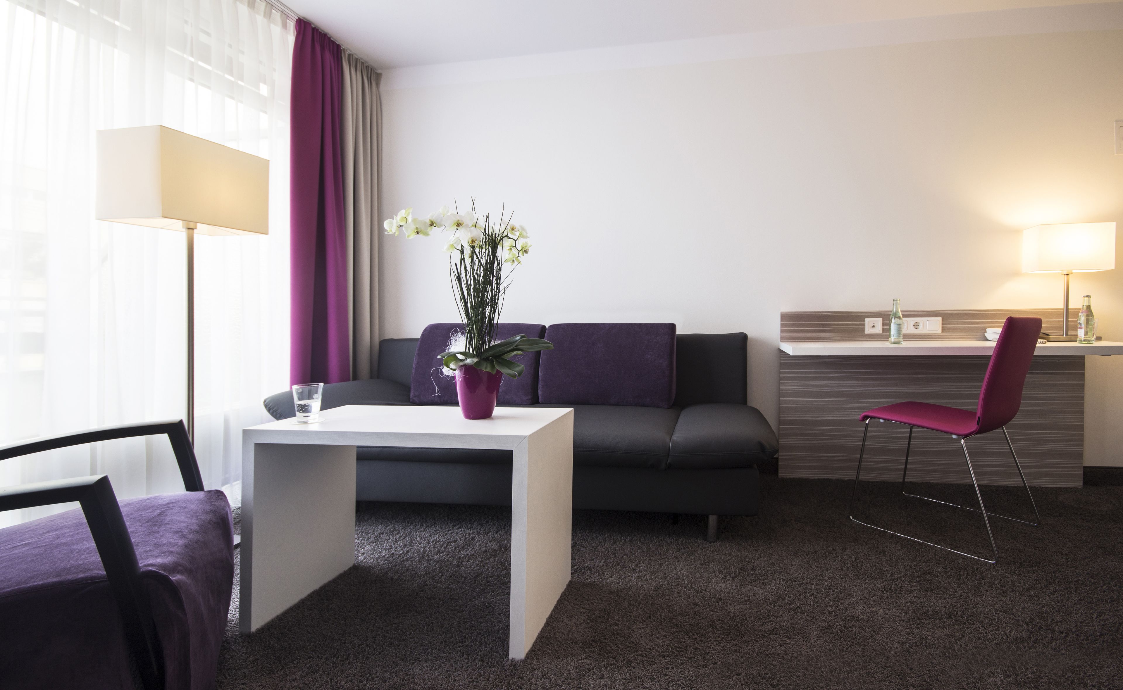A modern hotel room with a grey sofa, a white coffee table, a purple armchair, and a small dining area with a red chair.