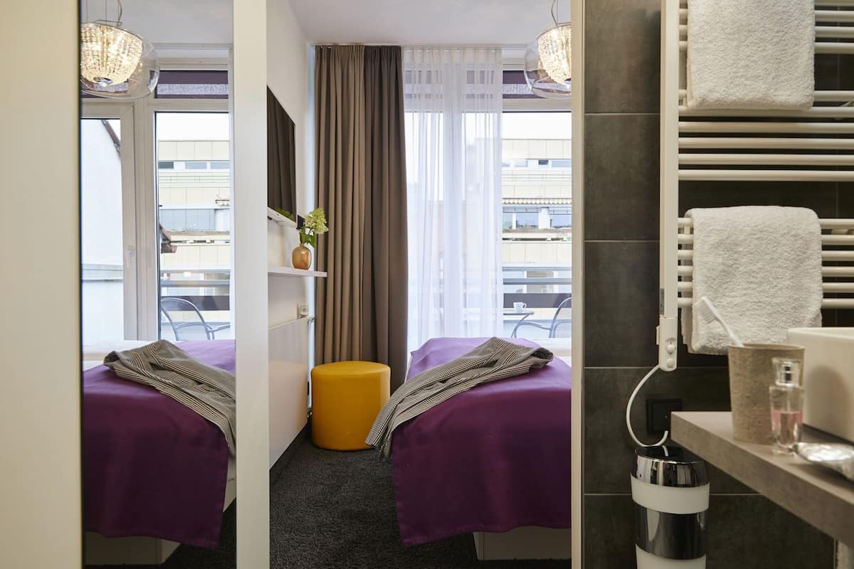 Camera d'albergo con due letti, un tavolino, una sedia, una finestra con tende e bagno con portasciugamani e lavandino.