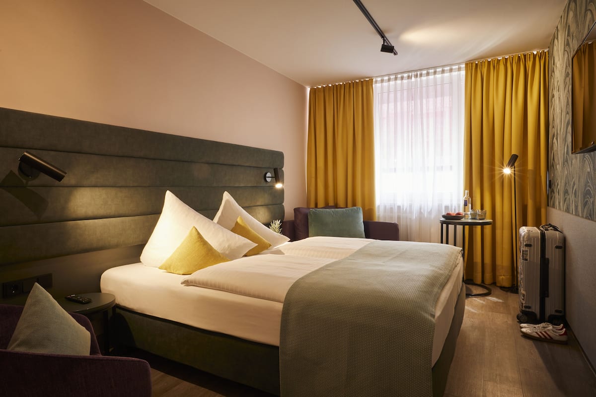 Una camera d'albergo con un letto grande, un comodino, una lampada, una sedia e una valigia.