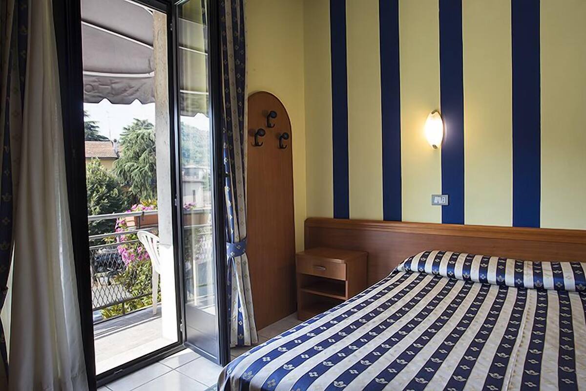 A bedroom with a bed, a wooden headboard, a nightstand, a wall-mounted light, and a view of the outside balcony.