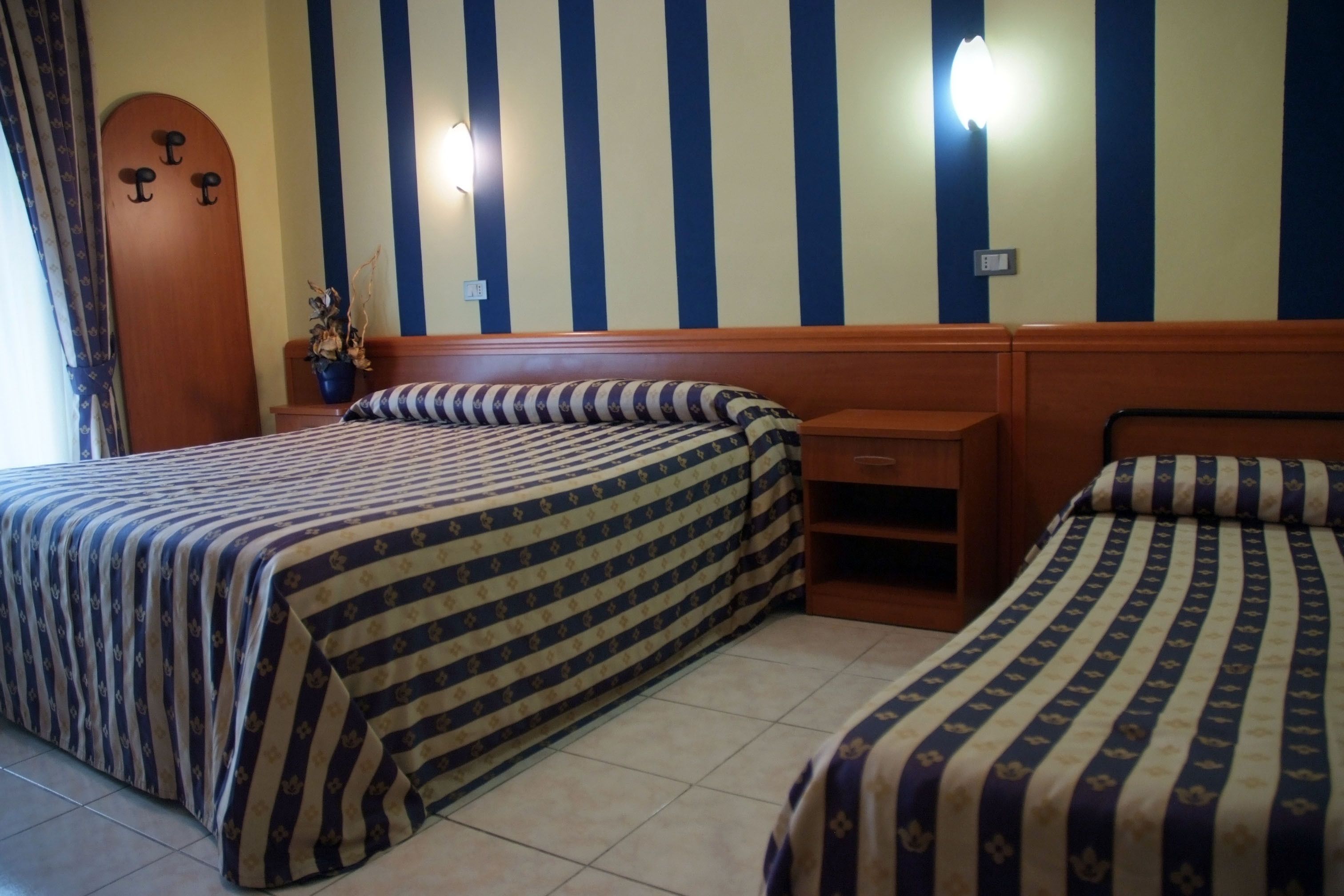 Une chambre d’hôtel avec deux lits, une literie à rayures, une tête de lit en bois, une table de chevet et une lampe fixée au mur.