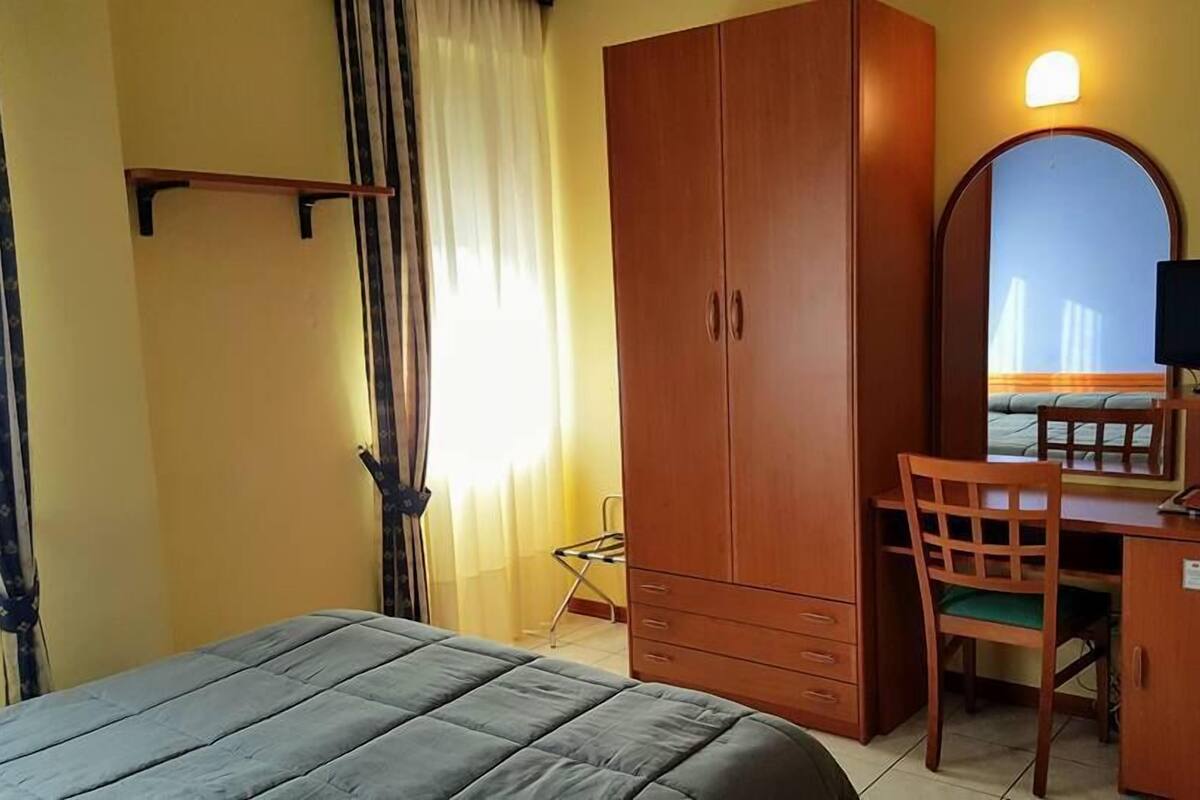 A hotel room with a bed, a desk with a chair, a wardrobe, a television, and a window with curtains.