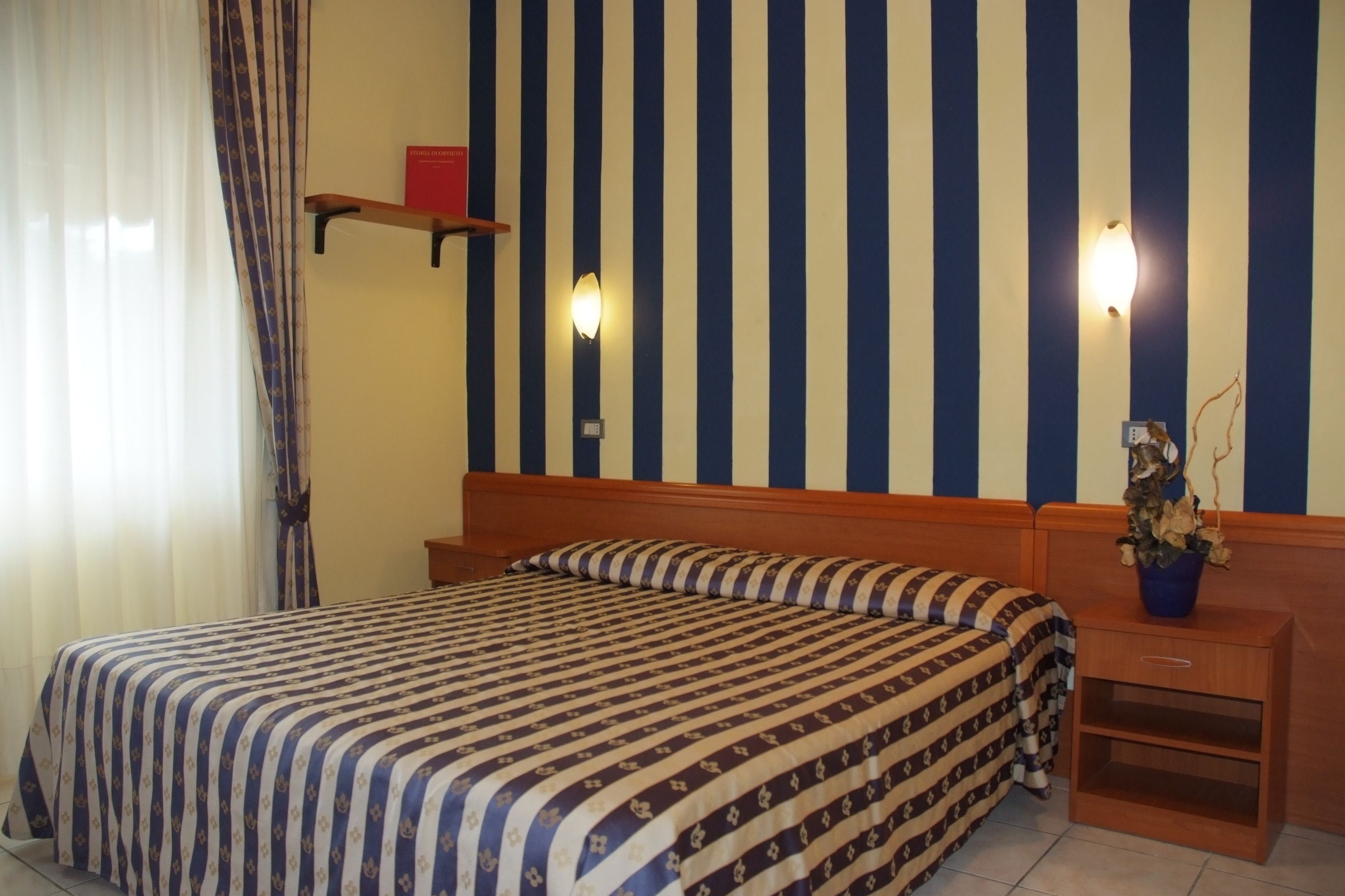 A hotel room with a striped wall, a bed with a patterned bedspread, a nightstand with a plant, and a shelf with a book.