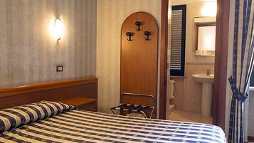 Una camera d'albergo con un letto, una porta in legno con ganci, un bagno con lavandino e specchio, e una finestra con tende.