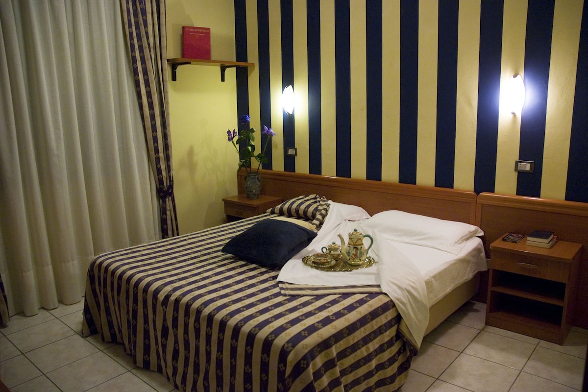 A hotel room with a bed, a nightstand, a bedside table, a vase with flowers, and a striped wall.