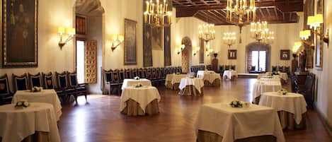 A spacious banquet hall with multiple round tables covered with white tablecloths, wooden chairs, and ornate chandeliers.