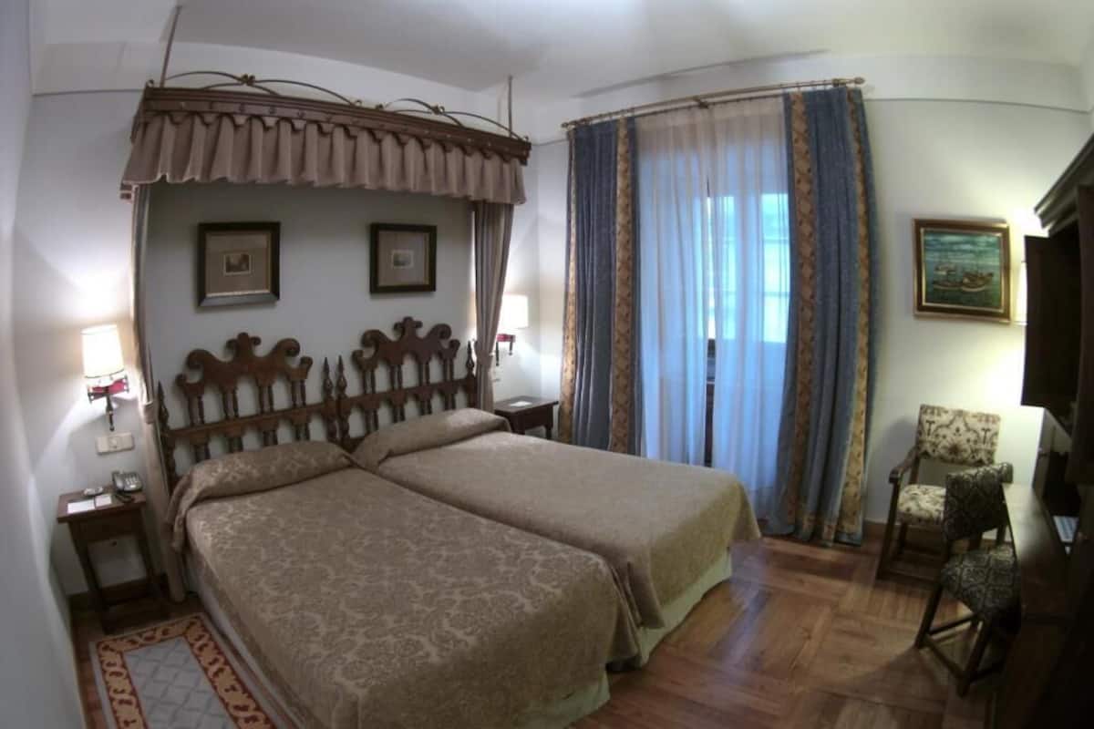 Un lit double avec une tête de lit, une table de chevet, une chaise, un bureau et une fenêtre avec des rideaux.