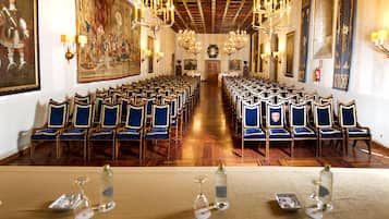 Um salão formal com fileiras de cadeiras estofadas em azul, uma mesa comprida com garrafas de água e copos, e um candelabro.